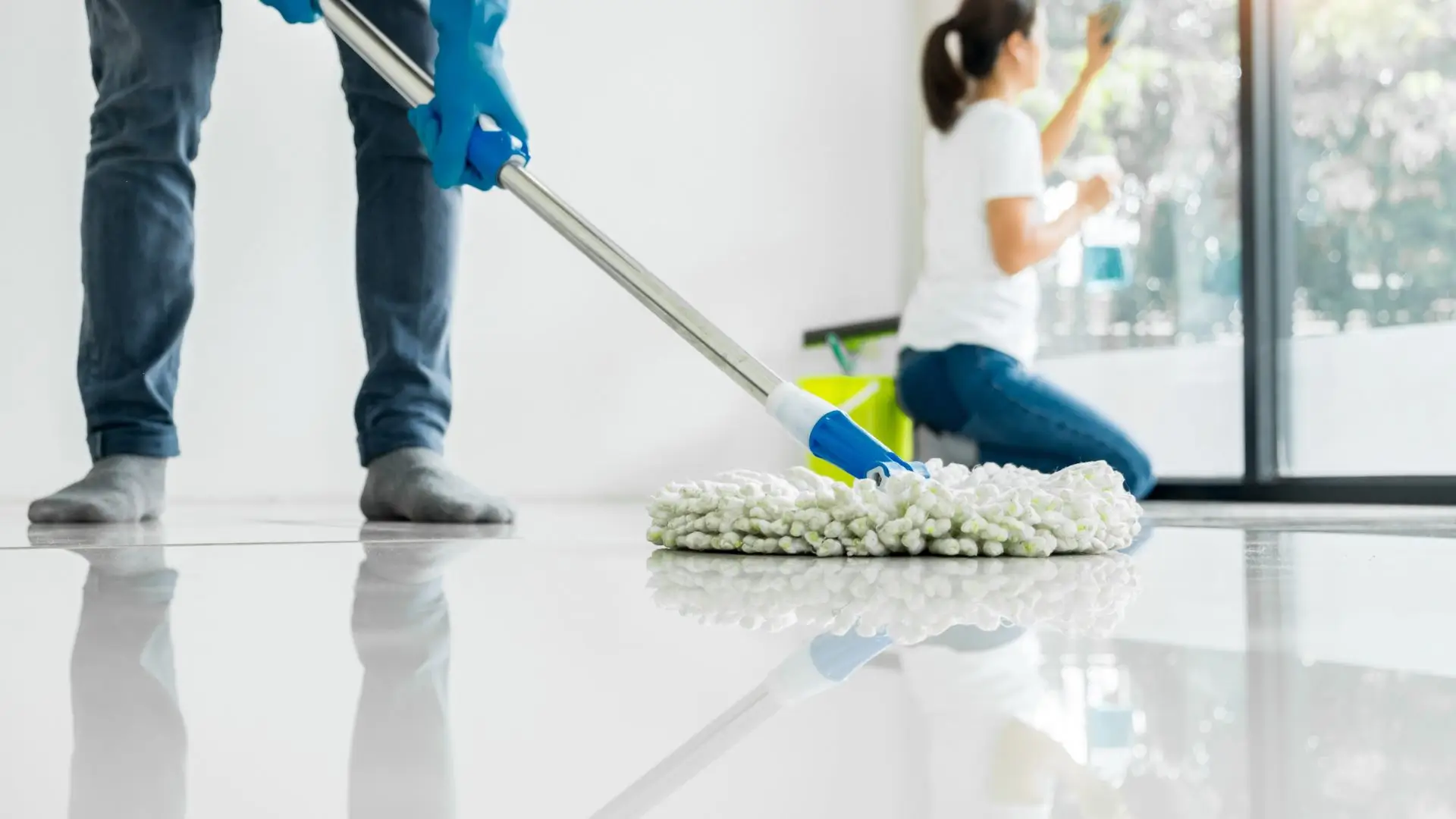 A professional cleaner mopping a tile floor while another team member cleans windows, ensuring a pristine home or office.